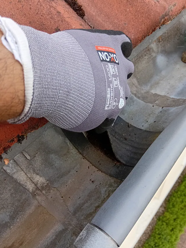 Hand im Arbeitshandschuh holt Laub und Schlamm aus einer Zinkrinne